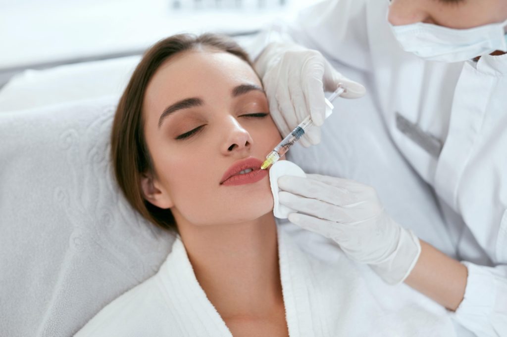Young woman receiving injection of filler in lips - Cosmetologist holding syringe and woman's face in hands Getting Filler treatment | Beauty at First Sight Med Spa in Chicago, Illinois